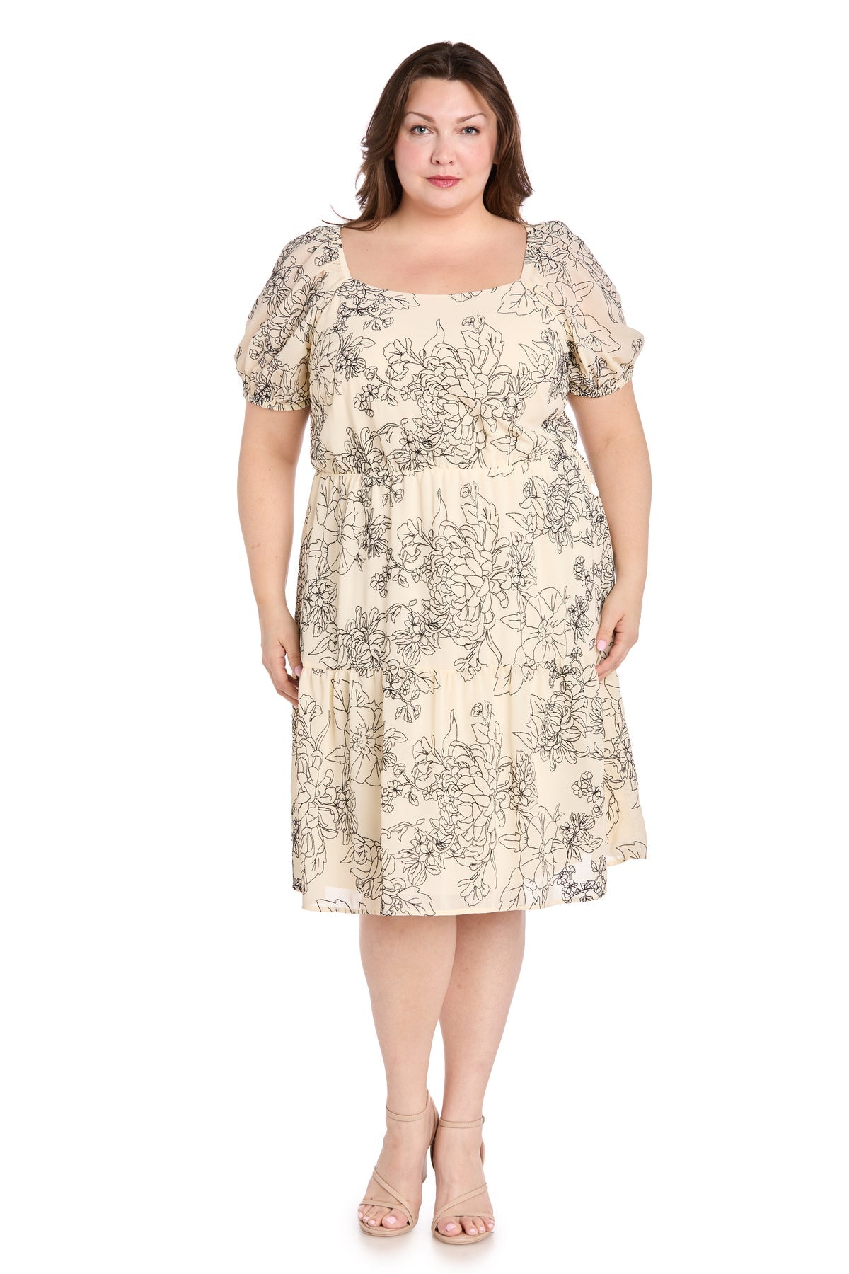 A woman faces forward in the Plus-Size Ivory Party Dress: R&M Richards 6688W, a knee-length ivory dress with black floral print and nude heels, shown against a plain white background—ideal as a floral print cocktail dress.